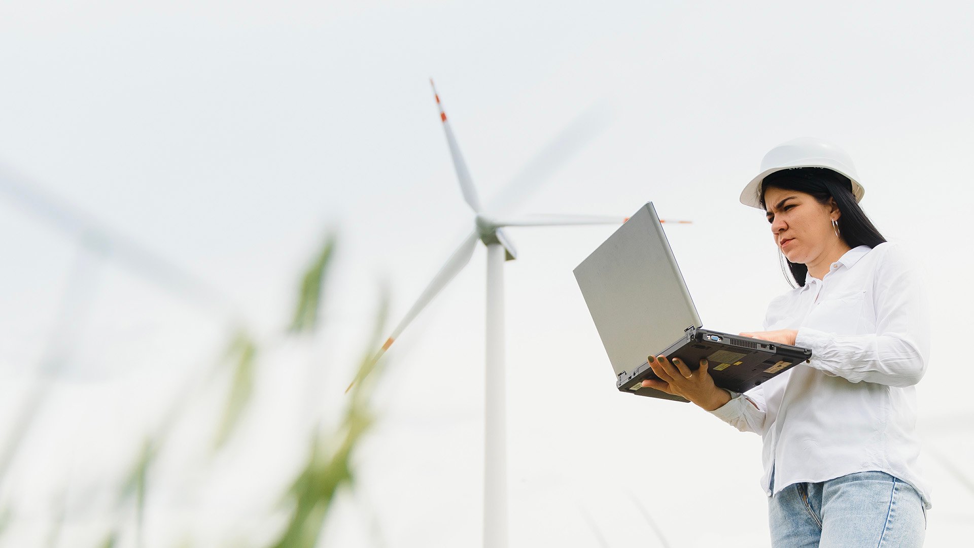 Wind technician in a hard hat working on a laptop near a wind turbine